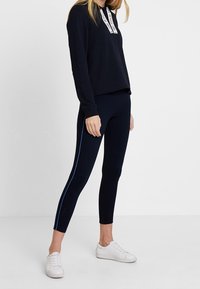 Marine blauwe hoodie met een witte logo-trekkoord, gecombineerd met marineblauwe leggings met een blauwe zijstreep, en witte sneakers. Glad oppervlak.