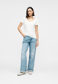 Λευκό βαμβακερό T-shirt με στρογγυλή λαιμόκοψη και ανάγλυφο λογότυπο, σε συνδυασμό με ανοιχτό μπλε τζιν παντελόνι φαρδιάς γραμμής με σκισίματα. Μπεζ παπούτσια slip-on.