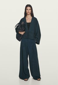 Tmavěmodrá, oversized, zkrácená bunda s širokými rukávy, kombinovaná s odpovídajícími kalhotami s vysokým pasem a širokými nohavicemi. Modelka nese černou tašku.