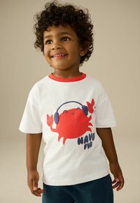 T-shirt en coton blanc avec des finitions rouges, représentant un crabe cartoon portant des écouteurs et le texte "AMUSEZ-VOUS" en bleu. Manches courtes, coupe décontractée.