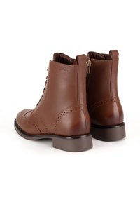 Hnedé kožené členkové topánky s brogue detailmi, šnurovací dizajn, bočný zips a blokový podpätok. Hladká textura s jemným brandingom.