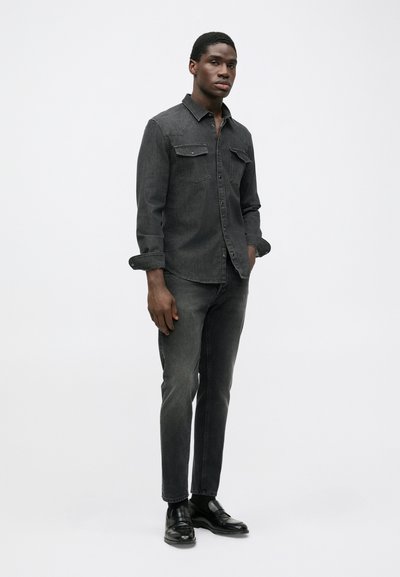 Dark denim shirt with two chest pockets, button-down front, rolled sleeves; paired with dark jeans and black shoes, creating a monochromatic look.