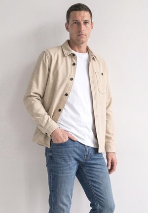 Mand med kort mørkt hår iført beige skjortejakke, hvid T-shirt og blå jeans, stående mod en ensfarvet lys væg.