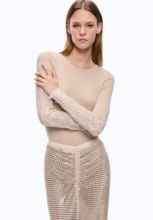 Mujer con el pelo liso, vestida con una camiseta beige acanalada con mangas de encaje y una falda beige texturizada, de pie con los brazos cruzados.