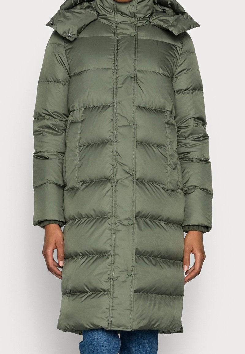 Long manteau d’hiver matelassé vert olive avec boutons-pression, poches latérales et capuche amovible, porté sur un jean bleu.