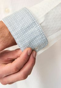 Poignet de chemise à manches longues présentant un motif géométrique bleu et blanc avec un tissu blanc texturé en dessous. Détails des boutons visibles.