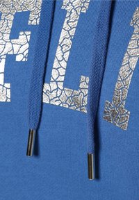 Sudadera azul con un destacado detalle de letras plateadas agrietadas y cordones gruesos con puntas metálicas. Tejido suave y texturizado.