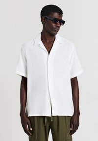 Chemise blanche à manches courtes avec un tissu texturé et une coupe décontractée, associée à un pantalon cargo vert olive. Lunettes de soleil noires portées.
