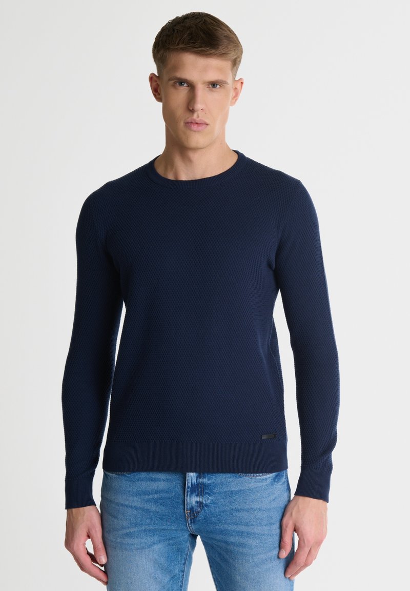 Temno moder, teksturiran pleten š sweater s okroglim izrezom in dolgimi rokavi, z ribanim robom in subtilno logotipno podrobnostjo na dnu.