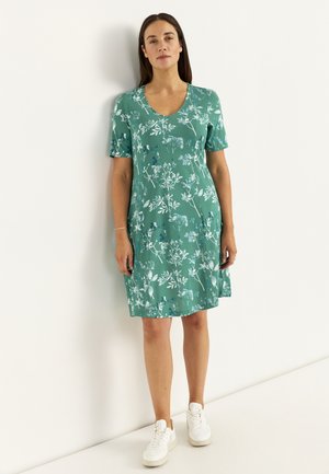 Kvinde står op ad en hvid væg iført en grøn kortærmet kjole med hvidt blomsterprint og hvide sneakers.