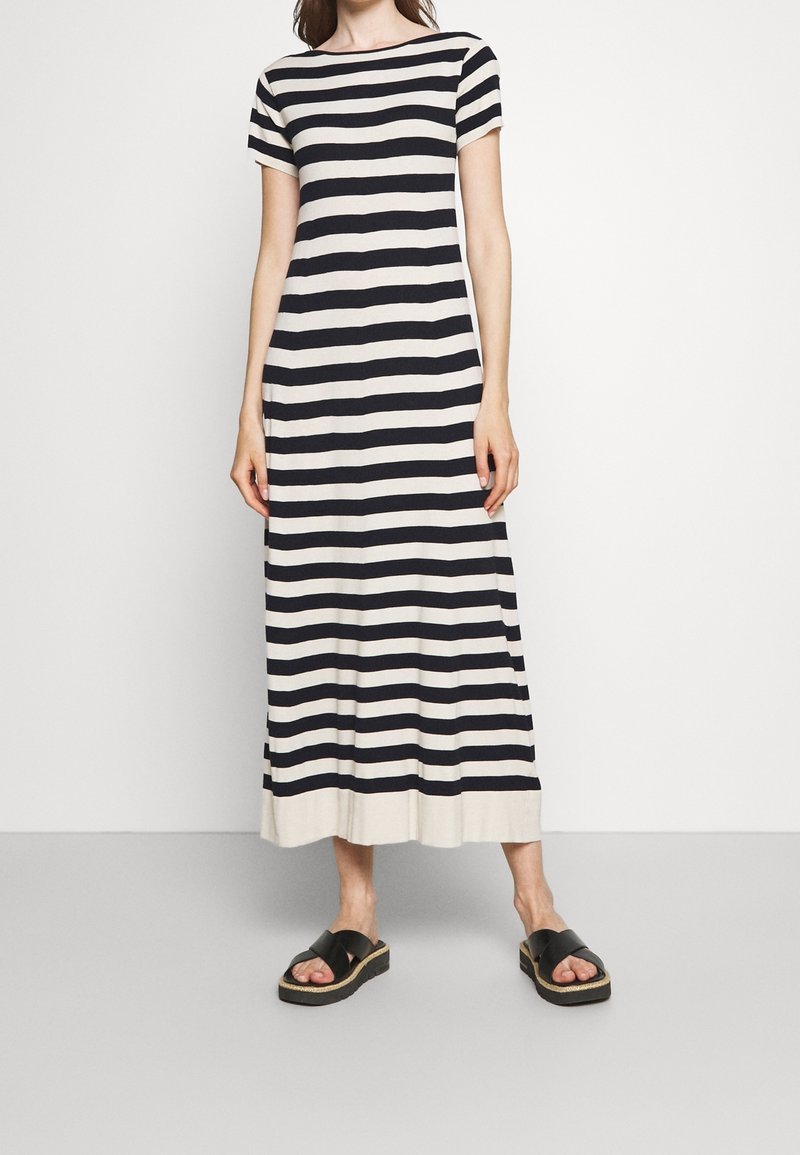Femme portant une robe maxi à manches courtes rayée horizontalement en noir et blanc avec des sandales noires, debout devant un fond blanc uni.