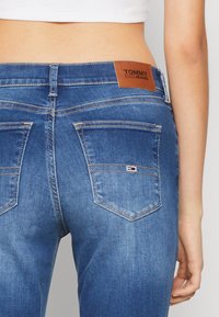 Jeans de denim azul con un diseño ajustado, que presentan una etiqueta de cintura de cuero marrón y dos bolsillos traseros con sutiles detalles de costura.