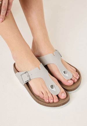 Grijze leren teenslippers hebben een uniek ontwerp met een teenlus, een zilveren gesp en een bruin gemarmerd voetbed met een witte zool.