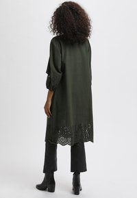 Vestido túnica verde oliva con dobladillo cortado con láser e mangas abullonadas; combinado con pantalones negros y botas de tobillo; textura suave.