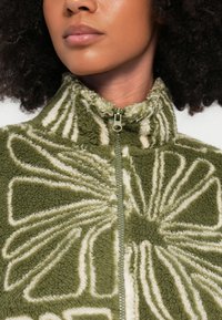 Chaqueta de forro polar verde con patrón floral blanco, cuello alto y cierre de cremallera. La tela texturizada crea una apariencia suave y acogedora.