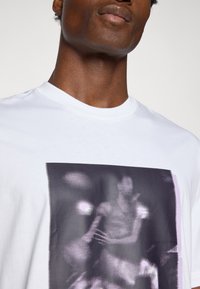 White cotton t-shirt with a graphic print featuring a pixelated basketball player in black and purple tones, round neckline, and short sleeves.