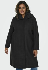 Manteau noir à capuche avec une silhouette droite, des manches longues et des poches latérales, fabriqué dans un tissu lisse au fini mat.