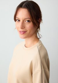 Beżowy sweter z okrągłym dekoltem i teksturowanymi srebrnymi akcentami na ramionach. Miękka tkanina o luźnym kroju. Gładka faktura.