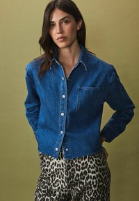 Een donkerblauwe denimshirt met knoopsluiting en een enkele borstzak, gecombineerd met broek in luipaardprint, tegen een olijfgroen achtergrond.