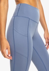 Leggings bleus taille haute en tissu extensible, avec des coutures tonales et une poche latérale pour le rangement. Texture douce.