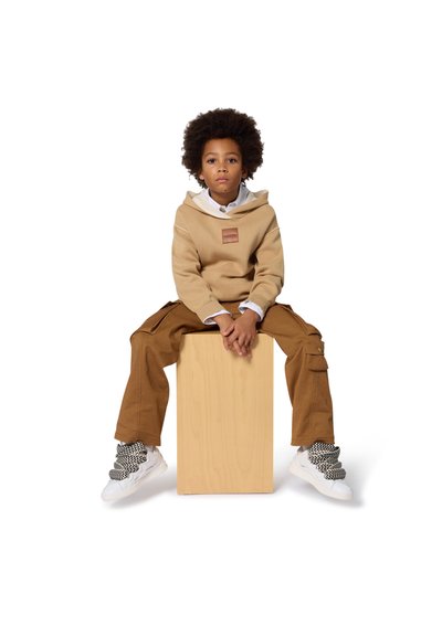 Felpa beige con un'etichetta in pelle, camicia bianca con colletto e pantaloni cargo marroni, seduta su un blocco di legno, completata da sneaker con fantasia.