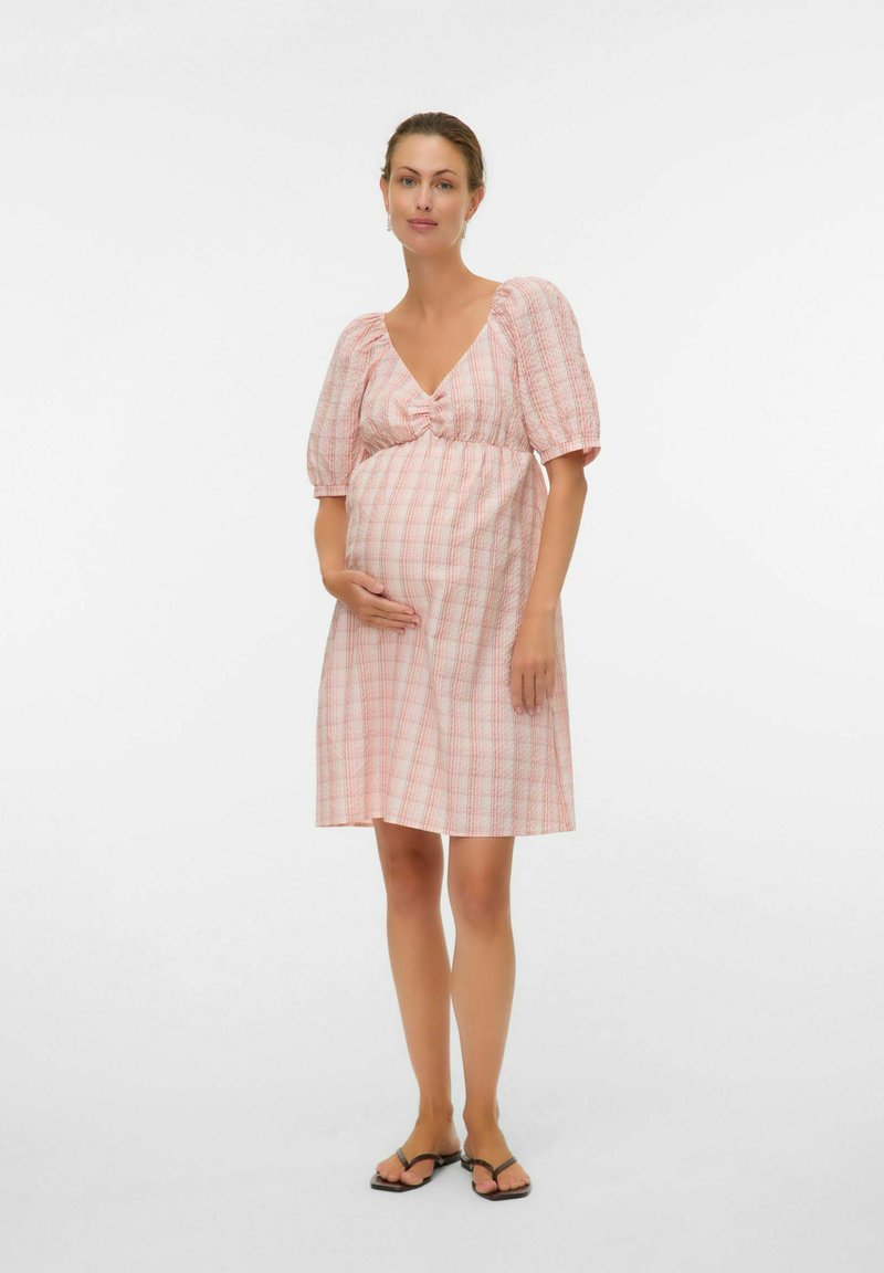 Femme enceinte debout, portant une robe à carreaux rose et blanche jusqu'aux genoux avec des manches bouffantes et des sandales noires, sur un fond blanc uni.