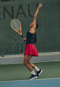 Sportoló teniszpályán, fekete ujjatlan pólót és piros rakott szoknyát viselve. Egy narancssárga teniszütőt tart, fekete markolattal.