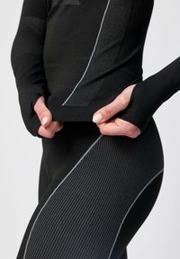 Svart aktivklädesset med långa ärmar och kroppsnära design. Har strukturerat ribbmönster och grå detaljerad söm. Nära bild som visar handens placering.