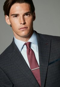 Jachetă de costum în carouri bleumarin, cu o cămașă albastră deschisă și o cravată bordeaux cu un model texturat și bară pentru cravată argintie. Design clasic al reverului.