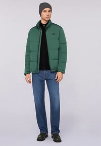 Zelená prošívaná bunda s vysokým límcem, zapínáním na zip a zatepleným designem, doplněná černým tričkem, modrými džínami a šedou pletenou čepicí.