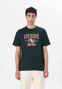 Camiseta de algodón verde oscuro con un gran logo rojo de "GUESS" y detalles en dorado, con cuello redondo y mangas cortas. Textura suave.