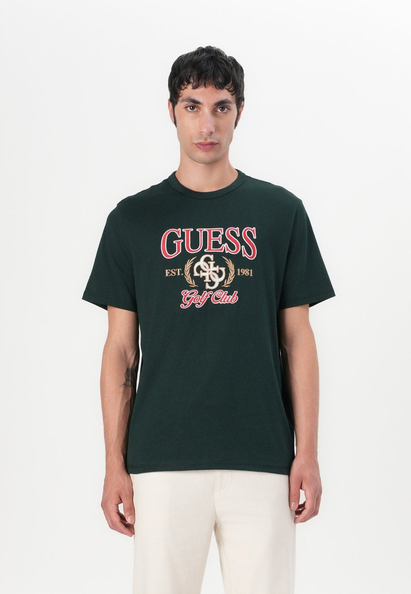 Camiseta de algodón verde oscuro con un gran logo rojo de "GUESS" y detalles en dorado, con cuello redondo y mangas cortas. Textura suave.
