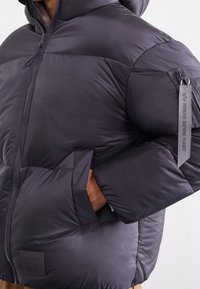 Dunkelgraue Pufferjacke mit Kapuze, ausgestattet mit einem Reißverschluss, gerippten Bündchen und einem kontrastierenden Etikett am Arm. Glattes, isoliertes Material.