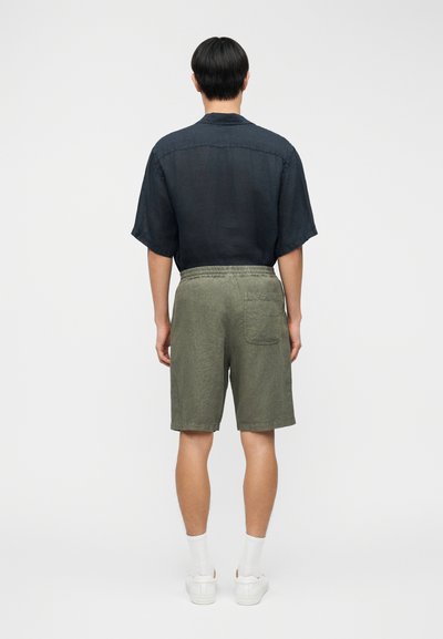 Chemise en lin bleu marine à manches courtes, associée à un short vert olive avec une ceinture élastique et une poche arrière. Des baskets blanches complètent la tenue.