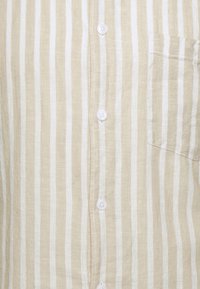 Camicia a righe beige e bianca in lino, con chiusura a bottoni sul davanti, taschino a toppa e texture liscia con cuciture visibili.