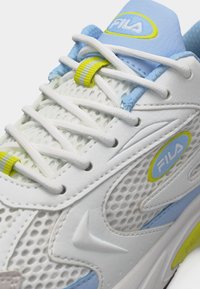 Λευκό και ανοιχτό μπλε αθλητικό παπούτσι FILA με υφή από δίχτυ, λευκά κορδόνια και κίτρινες ριγωτές θηλιές κορδονιών στη γλώσσα και την περιοχή του δακτύλου.