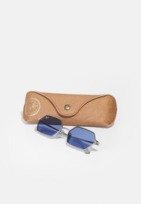 Ochelari de soare Ray-Ban cu lentile hexagonale albastre, rame metalice argintii și accente transparente, alături de un toc din piele maro deschis cu un logo auriu.