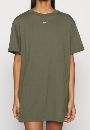Person trägt ein übergroßes olivgrünes Nike-T-Shirt mit einem kleinen weißen Swoosh-Logo, das in der Nähe des Halsausschnitts zentriert ist.