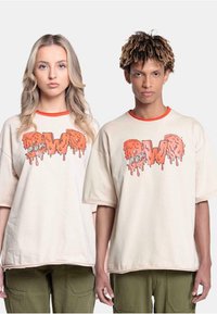 Beige oversized t-skjorter med oransje dryppgrafikk som sier "EWO," med korte ermer og kontrastfarge på halsen. Modellens grønne cargo-bukser er synlige.