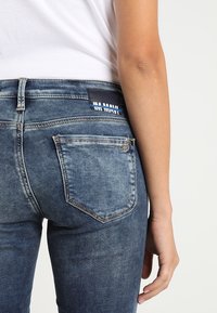 Smal blå denimjeans har hög midja, bakfickor och en svart etikett på midjebandet. Tyget har en sliten textur.
