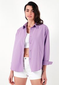 Lavender button-up shirt with a relaxed fit, long sleeves rolled up, and lightweight fabric. Paired with a white tank top and shorts.