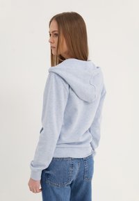 Sudadera azul claro hecha de material suave, con capucha ajustable, puños y dobladillo acanalados. Combinada con jeans de mezcla media.