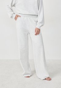 Person, der hellgraue, lockere Sweatpants und einen passenden Sweatshirt trägt, barfuß mit einer Hand in der Tasche vor einem schlichten Hintergrund steht.
