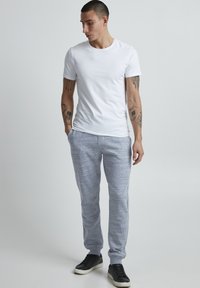 Hvit t-skjorte med korte ermer, kombinert med grå, teksturerte joggebukser og svarte sneakers, stående mot en ensfarget bakgrunn.