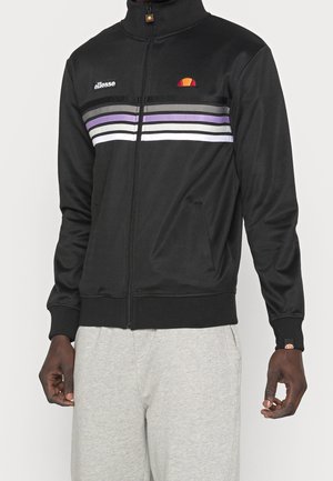 Czarna kurtka na zamek błyskawiczny z fioletowymi, białymi i szarymi pasami na wysokości klatki piersiowej, zestawiona z jasnoszarymi spodniami na stojącej osobie.