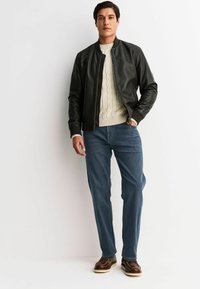 Schwarze Leder-Bomberjacke über einem cremefarbenen Kabelstrickpullover, kombiniert mit blauen Jeans und braunen Schuhen. Glatte Textur, figurbetontes Design.