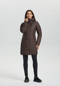 Brauner Puffer-Mantel mit Kapuze, Reißverschluss, horizontalen Nähten und seitlichen Taschen, kombiniert mit schwarzen Leggings und Turnschuhen.