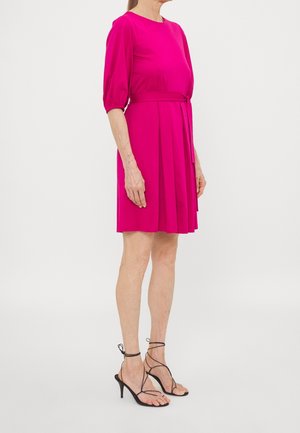 Mujer con vestido rosa brillante hasta la rodilla con mangas tres cuartos y sandalias negras de tacón con tiras, de pie de lado.