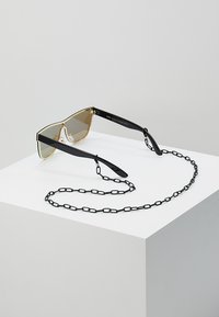 Saulesbrilles ar zelta metāla rāmi un brūnām tonētām lēcām, ar melnām kājiņām un noņemamu melnu ķēdi papildu drošībai.