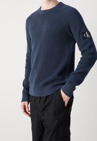 Sweatshirt en tricot bleu marine avec col rond côtelé, tissu texturé et un écusson noir sur la manche gauche ; associé à un pantalon noir.